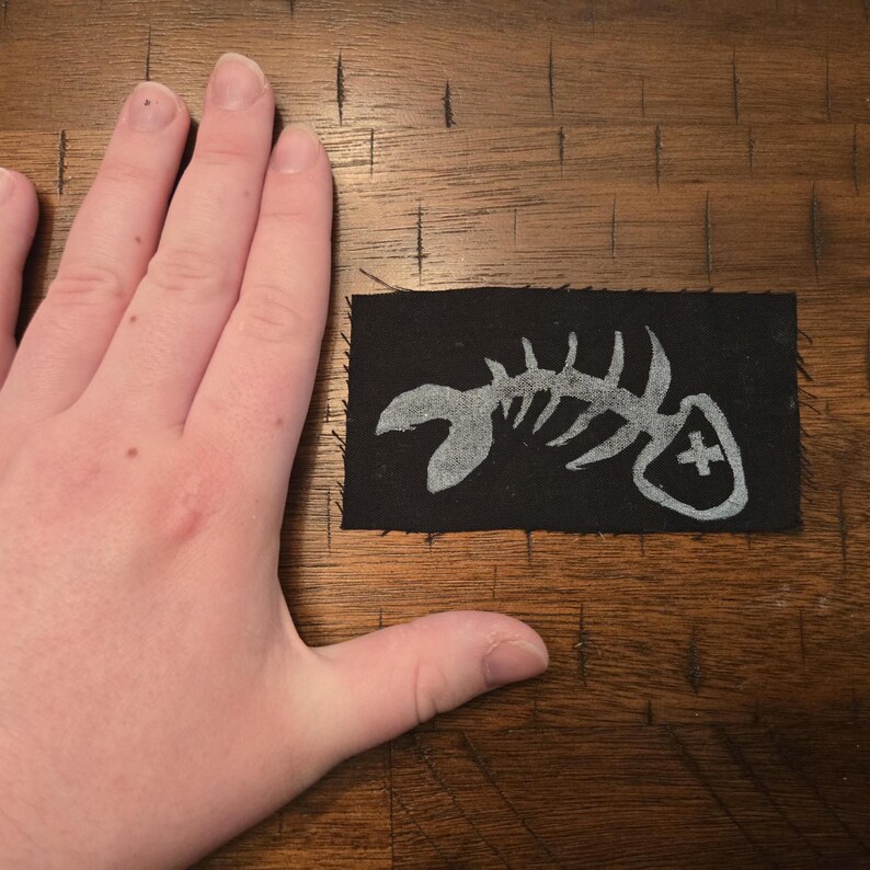 Pu&ograve; includere: Una toppa rettangolare nera con un disegno di scheletro di pesce argentato. La toppa ha un bordo cucito e un piccolo scudo con un simbolo a croce. La toppa &egrave; lunga circa 7,6 cm.