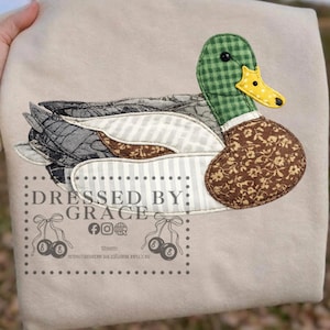 May include: A beige fabric item featuring an appliqued duck design. The duck has a green checkered head, a yellow beak, and a brown floral patterned body. The wings are a mix of gray, white striped, and brown patterned fabrics. The text "Dressed by Grace" is printed on the fabric.