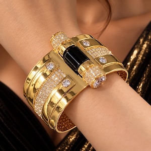 Peut inclure: Un large bracelet manchette doré avec un accent cylindrique noir central flanqué de deux sections en cristal pavé. La surface dorée polie est ornée de quatre cristaux ronds transparents et de rangées de petites pierres scintillantes.