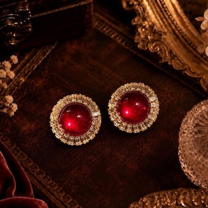 May include: A pair of round stud earrings featuring a large, smooth, deep red cabochon center stone surrounded by a halo of small, clear, rectangular-cut crystals set in a metallic frame that reflects light with a bright, polished finish.