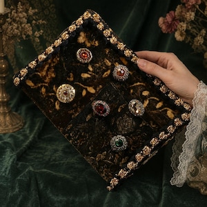Può includere: Un espositore di gioielli decorativo con un motivo floreale scuro e finiture dorate. La custodia contiene diversi anelli ornati con pietre preziose rosse, ambra e trasparenti. La custodia è tenuta da una persona con una manica di pizzo.