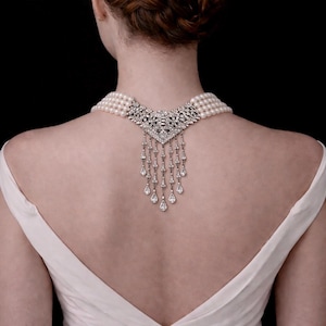 May include: A pearl necklace with a decorative silver pendant featuring a floral design and dangling teardrop-shaped gems. The necklace is worn on a person wearing a white dress, against a black background.