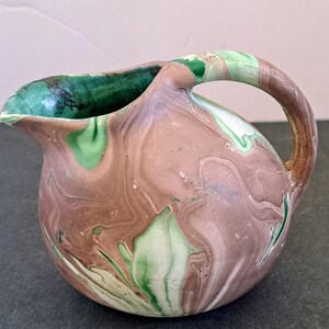 May include: A small, round ceramic jug with a spout and handle. The jug has a marbled pattern in brown, green, and white. The inside of the spout is dark green. The handle is curved. This jug is probably for serving liquids, such as milk or cream.