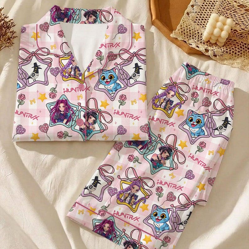 May include: Pink pajama set with a repeating pattern of cartoon characters, stars, and bows on a checkered background. The set includes a button-down shirt and matching pants. The word "HUNTRA" is printed on the pajamas.