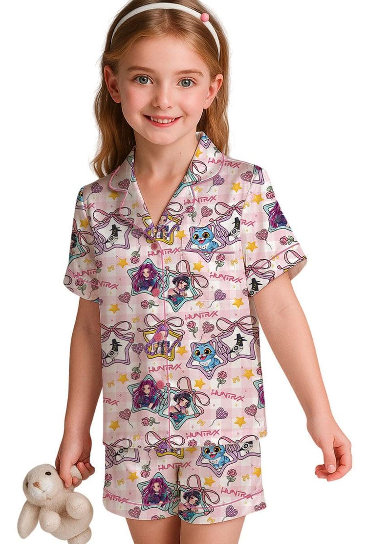 May include: Pink pajama set with a repeating pattern of cartoon characters, stars, and bows. The short-sleeved top and shorts feature the word "HUNTRIX" and are trimmed in pink. The set is made of a soft, silky material.