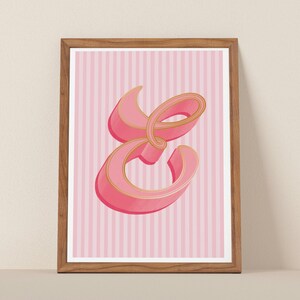 May include: A framed print featuring a stylized, three-dimensional letter E in shades of pink with gold-toned outlines. The letter is centered against a background of vertical light pink and white stripes, all enclosed in a simple wooden frame.