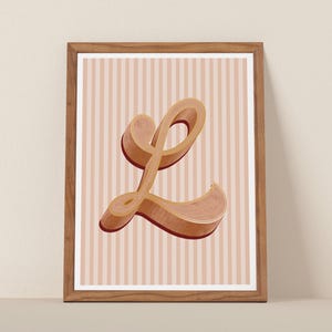 May include: A framed art print featuring a large, stylized letter "L" in a warm, three-dimensional design. The letter is a blend of brown and tan hues with a red outline, set against a background of vertical pink and white stripes. The frame is a natural wood color.