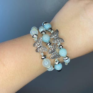 May include: A stack of beaded bracelets with silver butterfly charms. The bracelets feature light blue and white beads, with silver accents. The bracelets are worn on a wrist.