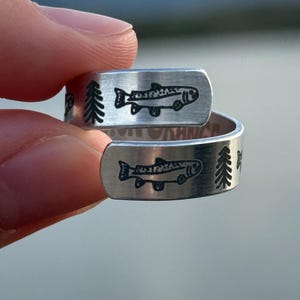 May include: Two silver rings with a fish and tree design. The rings are held up by a person's fingers. The rings have a brushed metal finish and a black etched design of a fish and trees.