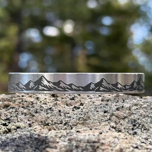May include: A silver-colored cuff bracelet with a mountain range design etched across the top. The mountains are depicted in black, creating a contrast against the metallic surface. The bracelet is resting on a textured rock surface.