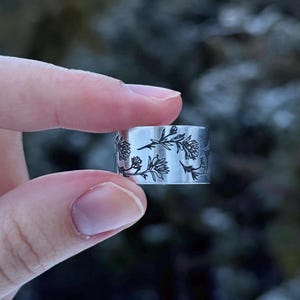 May include: A silver-colored ring with a floral design. The ring is held between two fingers. The floral pattern is etched in black around the band. The ring appears to be made of metal.
