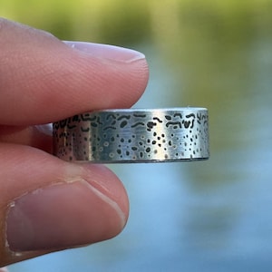 May include: A silver-toned metal ring featuring a flat band design with a textured surface. The exterior of the ring displays a pattern of dark, irregular, small dots and short horizontal markings that resemble a natural, organic speckled texture.