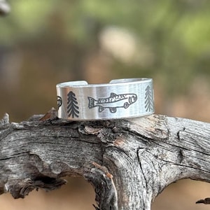 May include: A silver-colored adjustable ring with etched designs. The ring features a fish, and two trees. The ring is shown on a piece of weathered wood. The ring is a nature-themed accessory.
