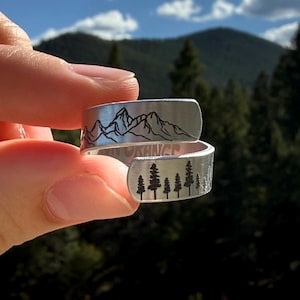May include: A silver-colored wrap ring with a mountain range design on one side and a forest scene on the other. The ring is held up against a backdrop of mountains and a blue sky.