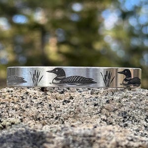 May include: A silver-colored cuff bracelet with a nature-inspired design. The bracelet features engraved images of loons and cattails. The loons are depicted in a detailed, black silhouette. The bracelet is displayed on a textured rock surface.