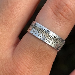 May include: A silver-toned band ring featuring a textured surface with a gradient pattern of small, dark, circular spots that transition from dense clusters to sparse markings across the metallic finish of the flat-profile ring band.