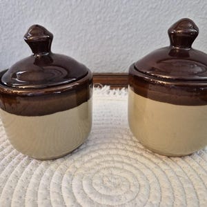 May include: Two matching ceramic jars with lids, each featuring a glossy dark brown glaze on the upper portion and a smooth, matte beige finish on the lower body. The lids have a rounded, knob-like handle at the top, and the jars are cylindrical.