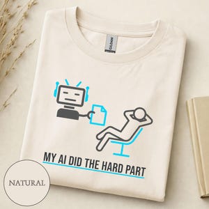 Könnte beinhalten: Cremefarbenes T-Shirt mit einer Grafik, die einen Roboter und eine Person zeigt, die sich in einem Stuhl entspannt. Der Text "MY AI DID THE HARD PART" ist unter der Illustration aufgedruckt. Das Shirt besteht aus einem weichen, natürlichen Stoff.