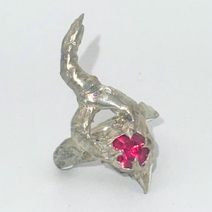 Op de afbeelding: Een zilverkleurige ring met een abstract, organisch ontwerp. De ring heeft een opvallend, gebogen hoornachtig element en een centrale cluster van helderrode edelstenen. De algehele stijl is artistiek en uniek.