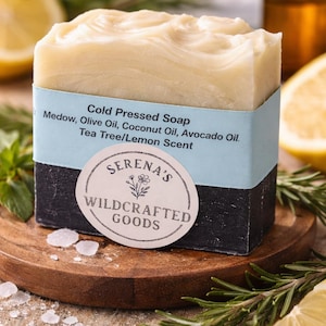 Könnte beinhalten: Ein rechteckiger Block kaltgepresster Seife mit hellbeigem Oberteil und dunkelgrauer Basis. Die Seife ist mit einem hellblauen Band mit Zutaten und einem runden Etikett mit der Aufschrift "Serena's Wildcrafted Goods" umwickelt. Zitronenscheiben und Rosmarinzweige umgeben die Seife.