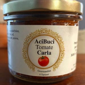 May include: A glass jar with a gold lid filled with a red spread. The label reads "AciBuci Tomate Carla" with a tomato illustration. Text on the label includes "Gourmandise à Tartiner".
