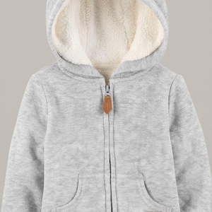 May include: A light gray zip-up hoodie featuring a hood with two small bear ears on top and a cream-colored sherpa lining. The jacket includes a front zipper with a brown leather-like pull, two front pockets, and a small brown patch with the text SMALL MIGHT.
