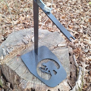 May include: A black metal stand featuring a cut-out silhouette of a person with a bow and arrow on its base. The stand has a vertical support and an angled arm with a bolt and two slots. It is on a tree stump.