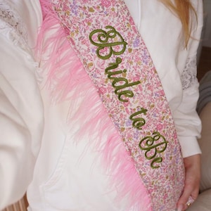 Könnte beinhalten: Eine Schärpe mit Blumenmuster in Rosa-, Lila- und Grüntönen, die am Rand mit rosa Federn besetzt ist. Der Text "Bride to Be" ist in grüner Schreibschrift mittig auf den Stoff der Schärpe gestickt.
