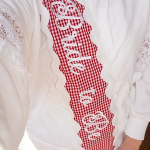 Puede incluir: Una sudadera con capucha blanca que presenta un panel vertical de cuadros vichy rojos y blancos en la parte delantera. El panel está bordeado por un ribete festoneado blanco y muestra el texto bordado en blanco "Bride to Be". Detalles de encaje blanco acentúan las mangas.