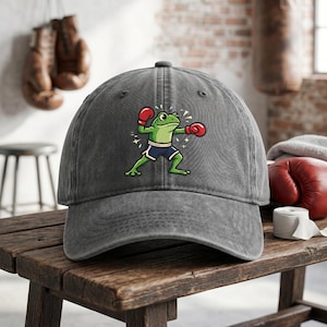 May include: A gray baseball cap featuring a cartoon frog in boxing gloves and shorts. The frog is green with red gloves and blue shorts. The cap is on a wooden surface.