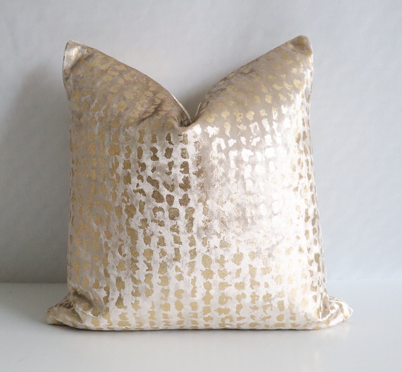 Puede incluir: Un coj&iacute;n decorativo cuadrado con fondo beige y un patr&oacute;n dorado. El patr&oacute;n dorado est&aacute; formado por peque&ntilde;as formas irregulares. El coj&iacute;n tiene una textura suave y aterciopelada, ideal para la decoraci&oacute;n del hogar.