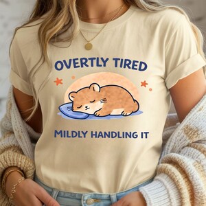 May include: Beige t-shirt featuring a cartoon hamster sleeping on a blue pillow, with the text "OVERTLY TIRED MILDLY HANDLING IT". The hamster is brown and the sun is orange.