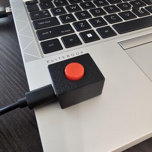 Könnte beinhalten: Ein quadratisches, schwarzes Makro-Tastengerät mit einem markanten, runden, orangefarbenen Knopf auf der Oberseite, das über ein schwarzes USB-Kabel mit einer silbernen Laptop-Tastatur mit der Aufschrift Elitebook verbunden ist.