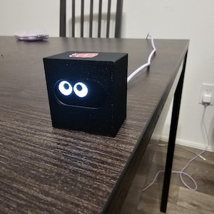 May include: A black, cube-shaped electronic device featuring a front-facing screen that displays two bright white, circular eyes. The device has a smooth, matte finish and is connected to a white power cable extending from the back of the unit.