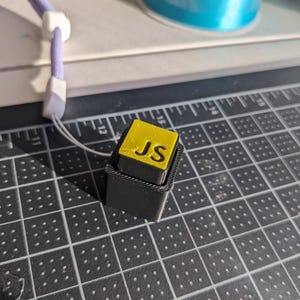 May include: A small, black, rectangular object with a yellow top. The top has the letters "JS" in black. A purple lanyard with a white clasp is attached to the object. A roll of blue ribbon is in the background.