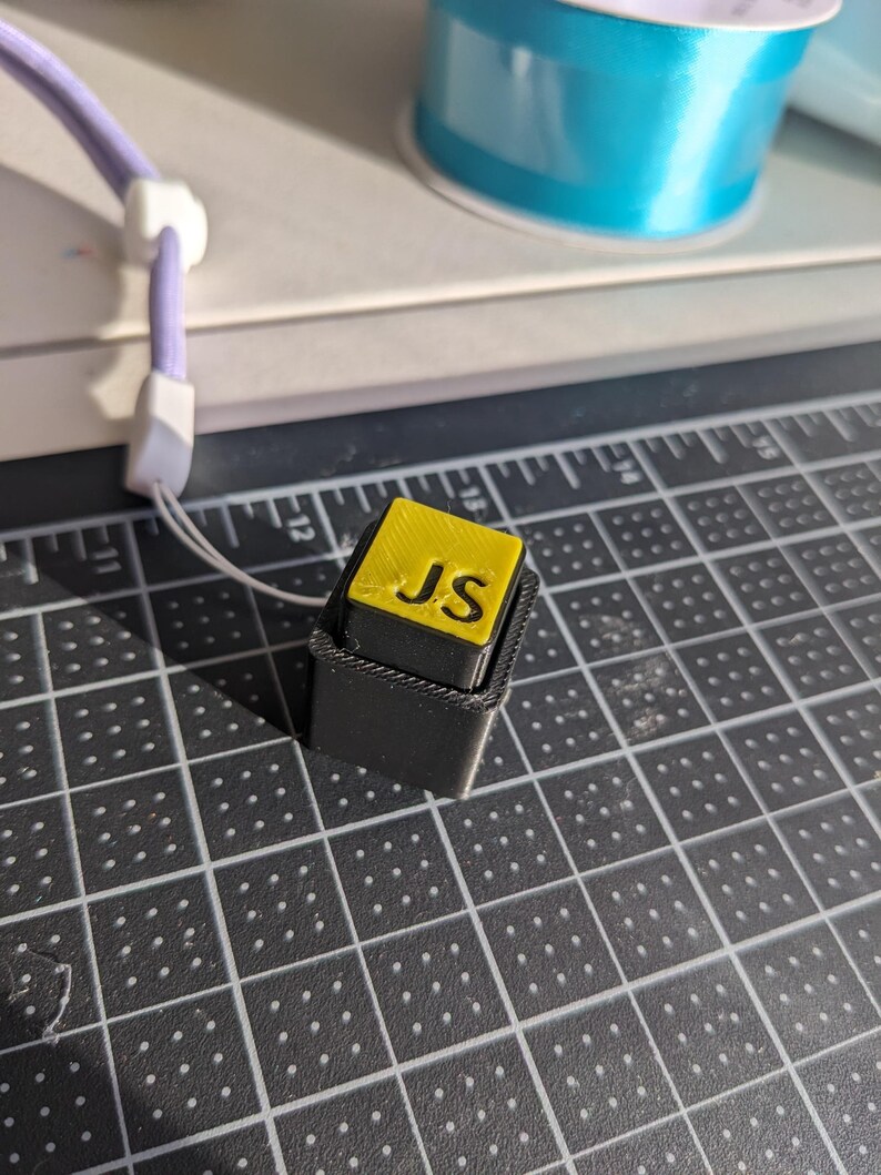 May include: A small, black, rectangular object with a yellow top. The top has the letters "JS" in black. A purple lanyard with a white clasp is attached to the object. A roll of blue ribbon is in the background.