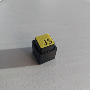 May include: A small, black, two-tiered block with a yellow top. The top tier features the yellow letters "JS". The object is sitting on a light grey surface.