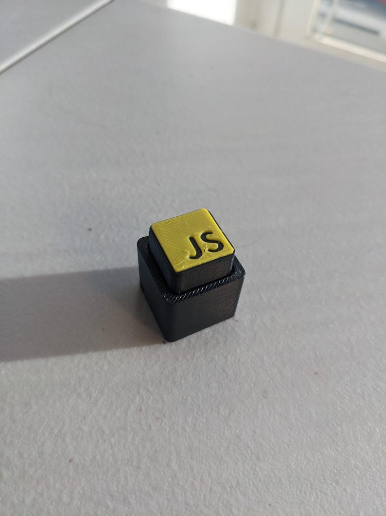 May include: A small, black, two-tiered block with a yellow top. The top tier features the yellow letters "JS". The object is sitting on a light grey surface.