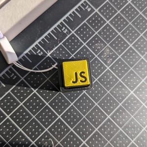 May include: A small, square, yellow and black object with the letters "JS" in black. The object is on a black grid surface with a white and purple lanyard attached. The letters are in a bold, sans-serif font.