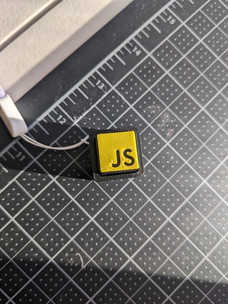 May include: A small, square, yellow and black object with the letters "JS" in black. The object is on a black grid surface with a white and purple lanyard attached. The letters are in a bold, sans-serif font.