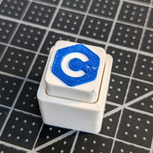 May include: A white, two-tiered block with a blue C symbol on top. The C symbol is a stylized, open-ended shape. The block is sitting on a black surface with a grid pattern.