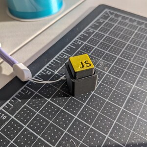 May include: A small, black, 3D-printed block with a yellow top. The top of the block has the letters "JS" in yellow. The block is on a black cutting mat with white grid lines and measurements in centimetres.