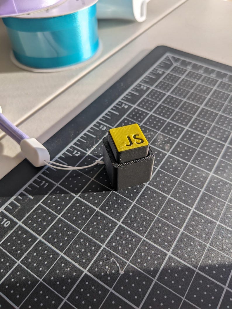 May include: A small, black, 3D-printed block with a yellow top. The top of the block has the letters "JS" in yellow. The block is on a black cutting mat with white grid lines and measurements in centimetres.