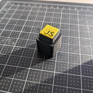 May include: A small, two-tiered, black and yellow block with the letters "JS" on the top. The top tier is yellow with black lettering, and the bottom tier is black. The object is sitting on a grey cutting mat.