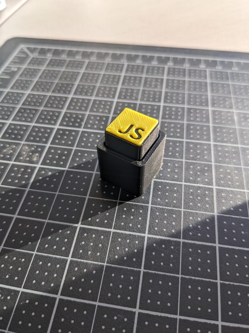 May include: A small, two-tiered, black and yellow block with the letters "JS" on the top. The top tier is yellow with black lettering, and the bottom tier is black. The object is sitting on a grey cutting mat.