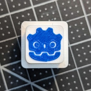 May include: A square button featuring a blue cartoon face design on a white background. The face has large eyes, a smiling mouth, and a crown-like shape. The button is likely for decorative or functional purposes.