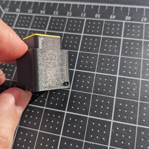 May include: A small, grey 3D-printed object with a yellow top is held between two fingers. The object has a rectangular shape with visible layer lines, indicating the printing process. The background is a black cutting mat with a grid pattern.