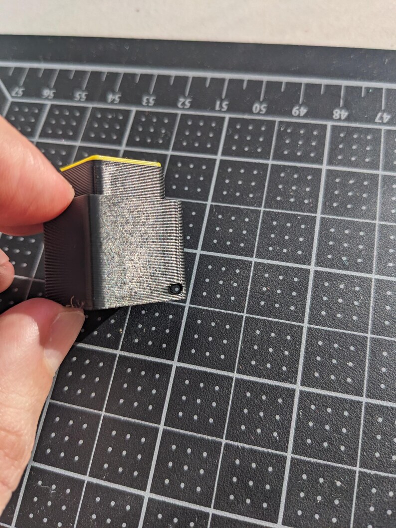May include: A small, grey 3D-printed object with a yellow top is held between two fingers. The object has a rectangular shape with visible layer lines, indicating the printing process. The background is a black cutting mat with a grid pattern.