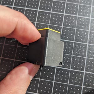 May include: A small, black, rectangular object with a yellow top. The object has a layered appearance and a small hole on one side. The object is being held by a person's fingers.