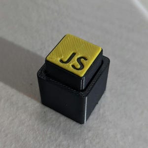 May include: A 3D-printed black and yellow square object. The top square is yellow with the letters "JS" cut out. The bottom square is black and slightly larger, creating a stepped effect. The object is likely a custom-made item.